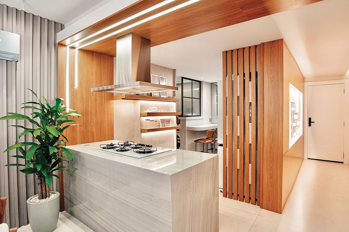 Cozinha e sala integradas em harmonia. Destaque para os painéis em tom Freijó, marcando a ilha e aquecendo o ambiente